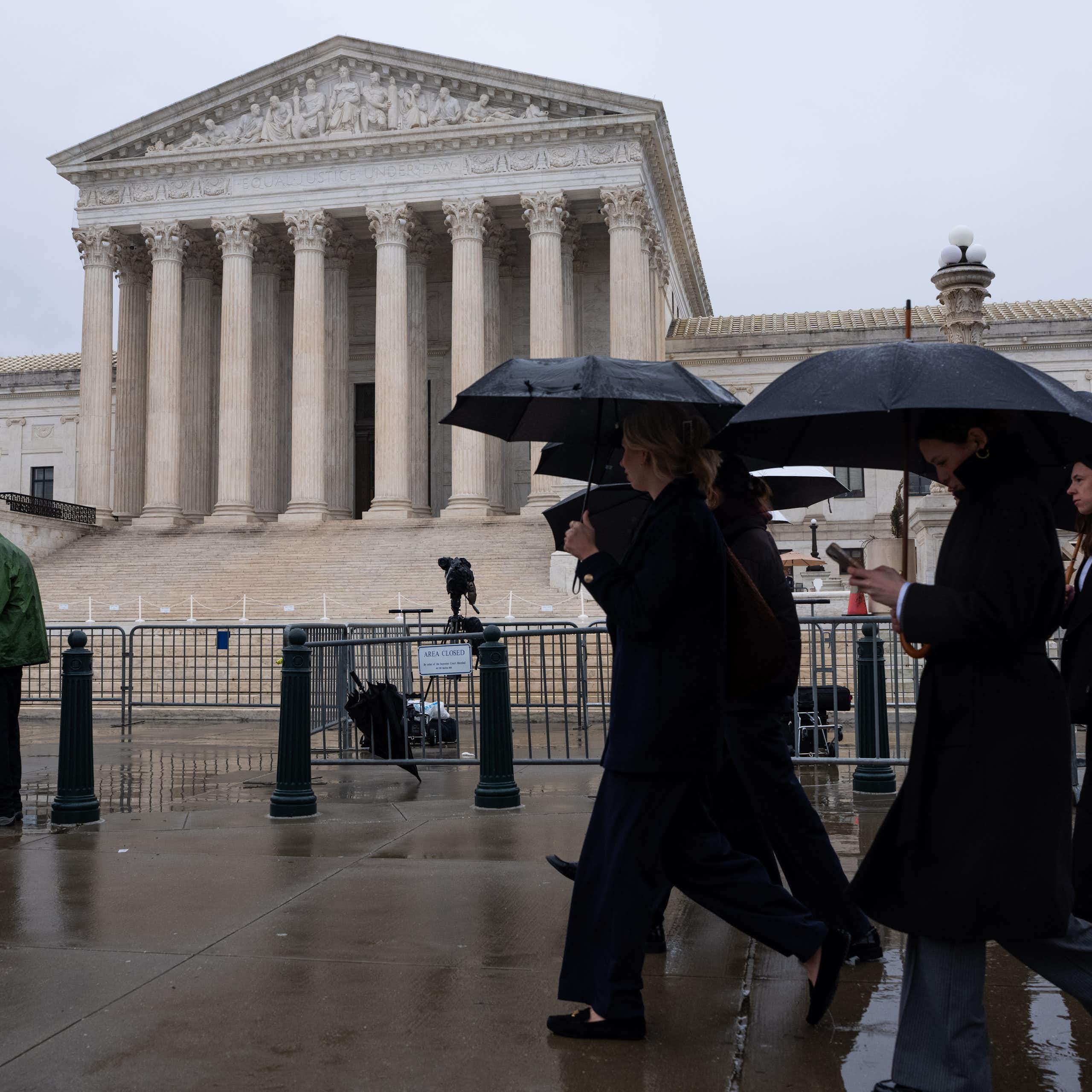 A group of people carry umbrellas as they walk past a big white building.