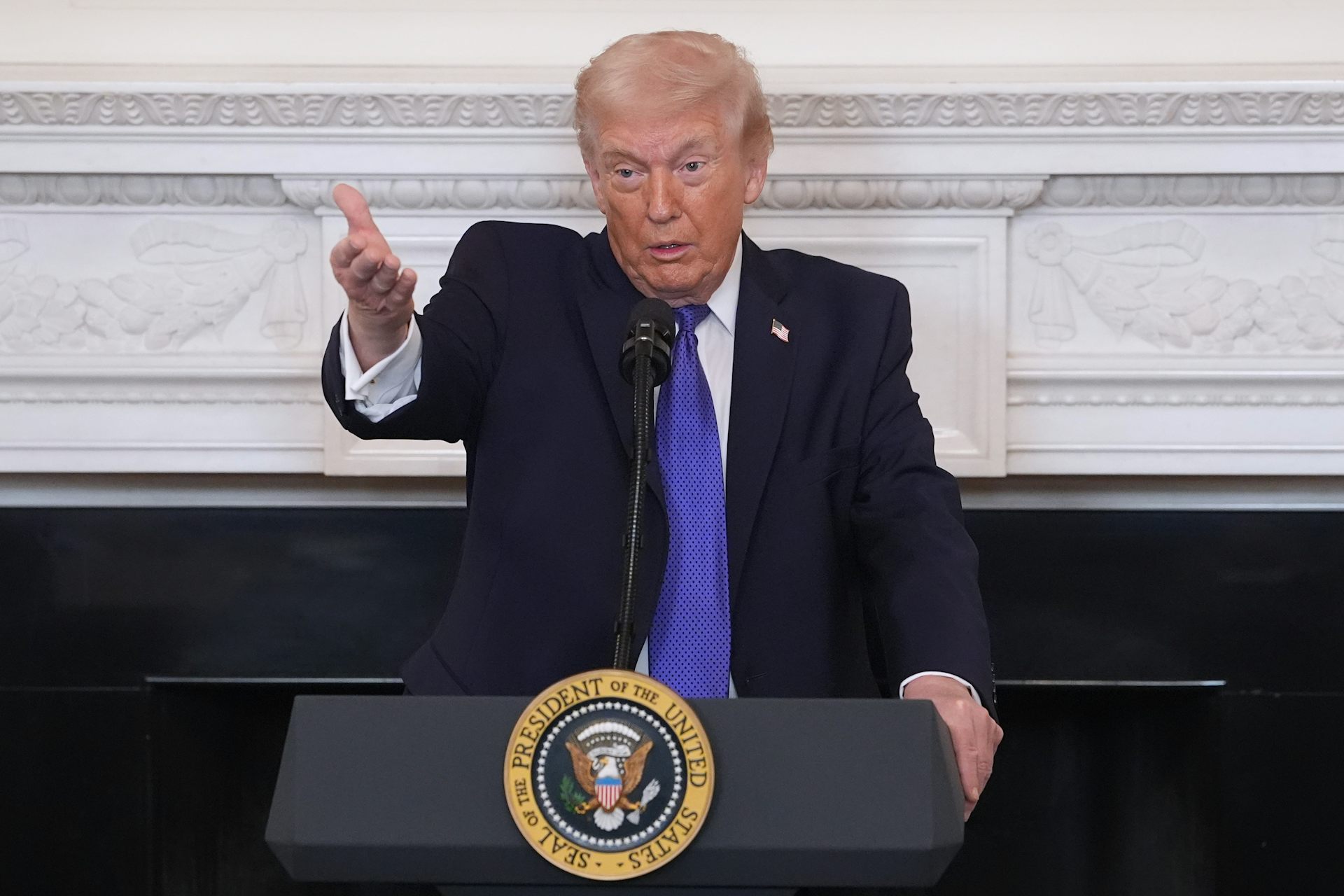 a white man in a suit gestures in front of a podium with the presidential seal on it