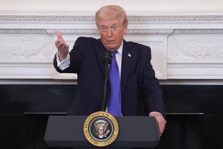 a white man in a suit gestures in front of a podium with the presidential seal on it