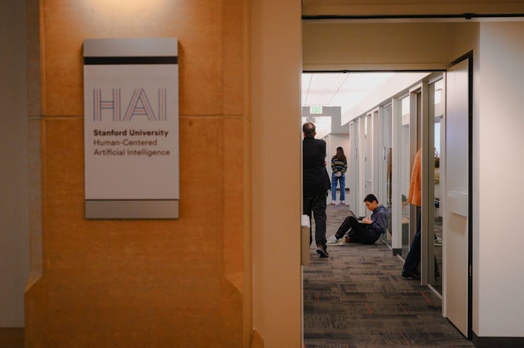 People are seen down a hallway, near a sign on a wooden wall that says 'Stanford University Human-Centered Artificial Intelligence.'