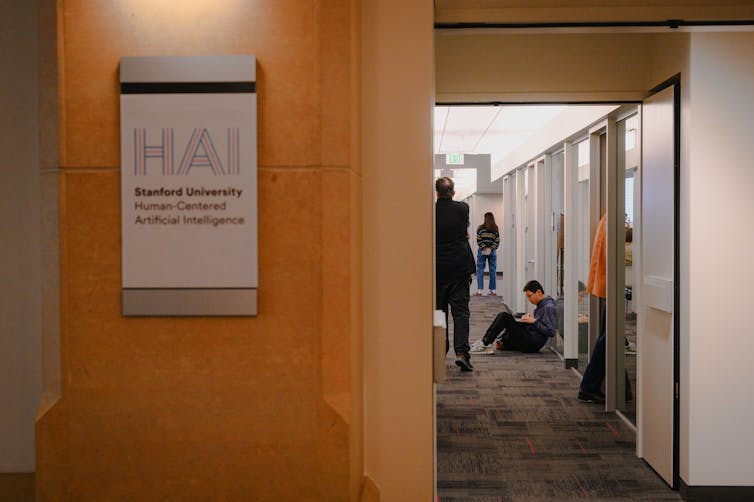 People are seen down a hallway, near a sign on a wooden wall that says 'Stanford University Human-Centered Artificial Intelligence.'