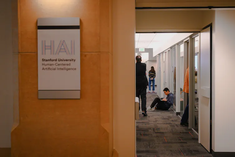 People are seen down a hallway, near a sign on a wooden wall that says 'Stanford University Human-Centered Artificial Intelligence.'