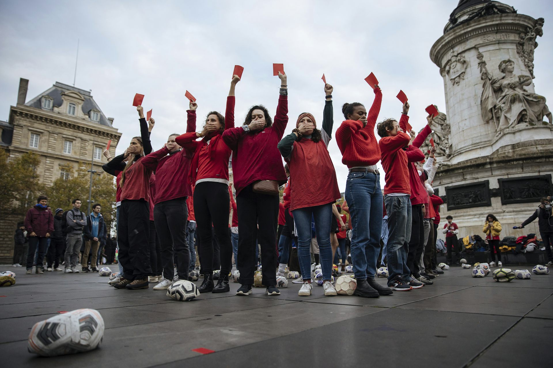 Un grupo de personas con camisetas rojas forman un círculo, tapándose la boca y sosteniendo tarjetas de fútbol rojas.