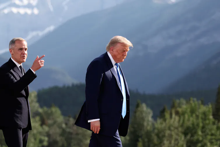 An old rotund orange-coloured white man in a dark suit. A thinner grey-haired man walks behind him pointing