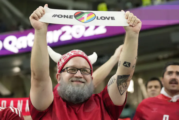 A bearded man in a red hat holds up a small banner that says 'one love'