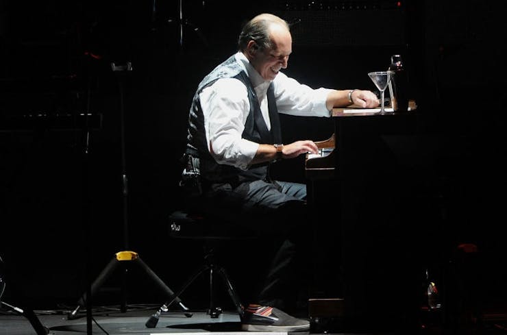 Balding, middle-aged man in a white shirt and black vest smiles while sitting at a piano on a stage.