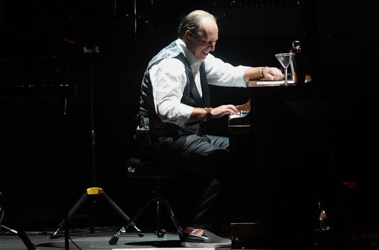 Balding, middle-aged man in a white shirt and black vest smiles while sitting at a piano on a stage.