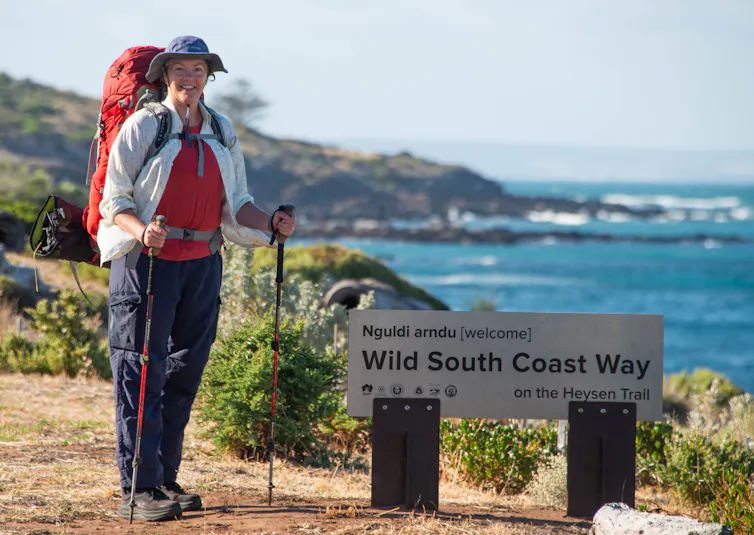 A woman standing by a sign that reads Wild South Coast Way, overlooking the sea