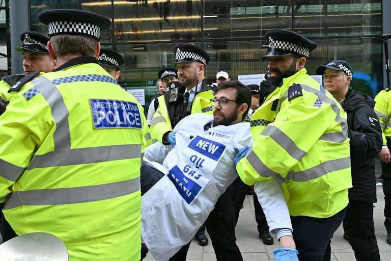 man in white lab coat being carried by police in outside protest