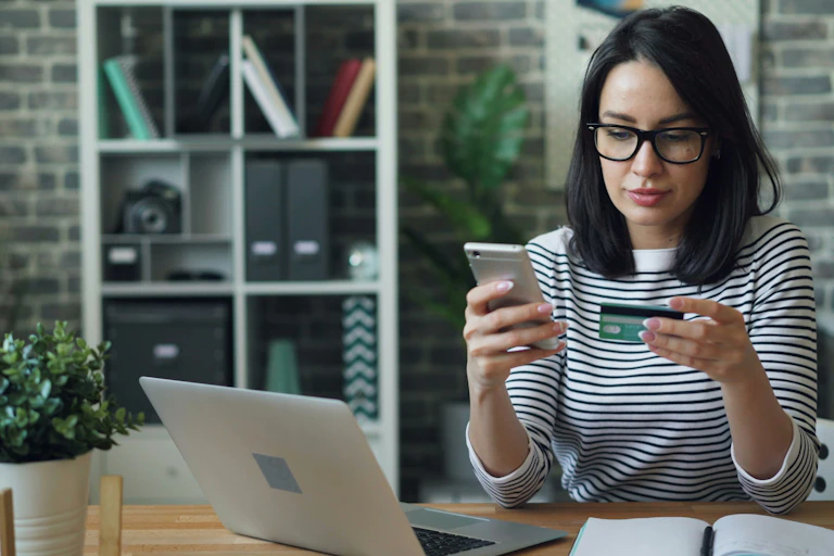 A woman looks at her credit card while typing on her cellphone