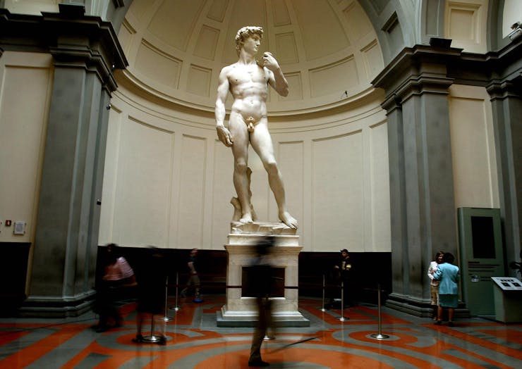 large white statue of nude man with blurry people passing its pedestal in museum