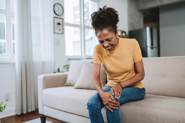 Person sitting on couch with hands clutching knees, grimacing in pain