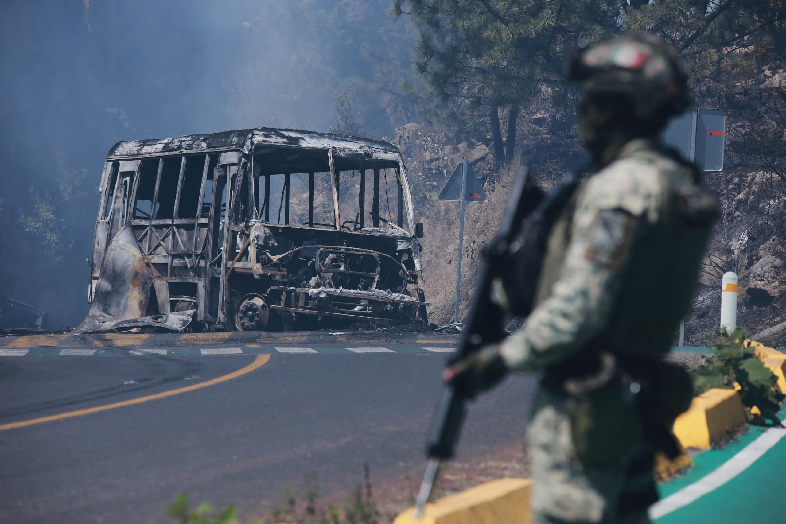 A man in fatigues carrying a gun stands in front of a charred vehicle.