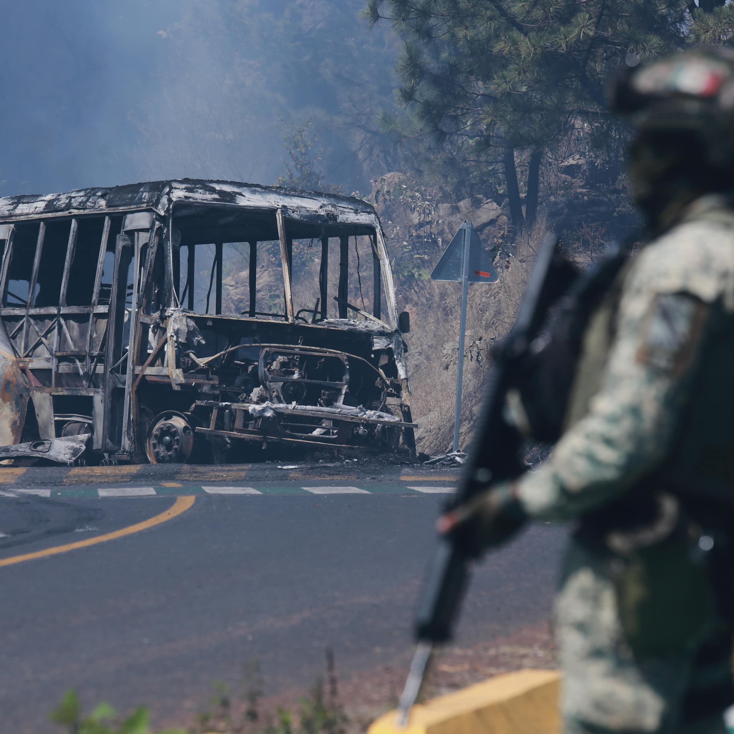 A man in fatigues carrying a gun stands in front of a charred vehicle.