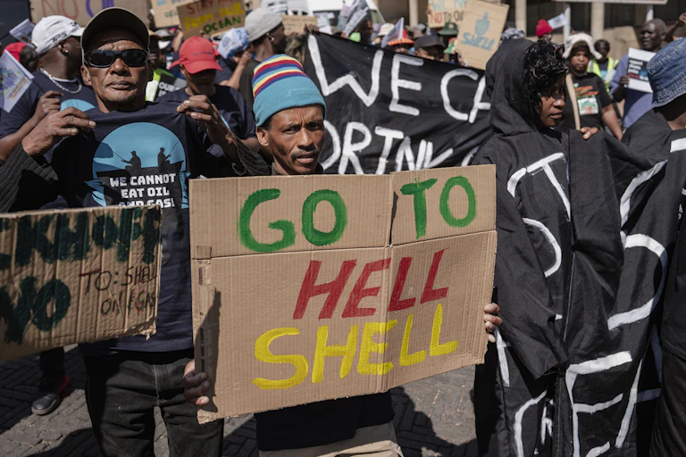Protestors marching in the street with a man in front holding up a placard that says 'Go to Hell, Shell'