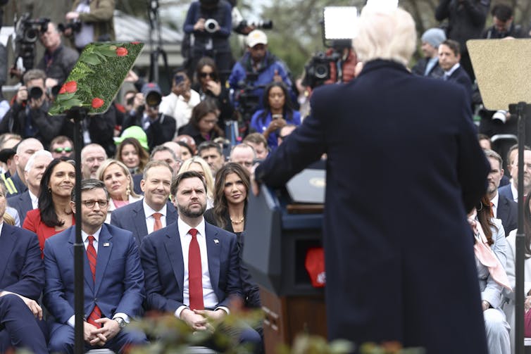rear view of donald trump at a lectern announcing liberation day tariffs with jd vance and others looking on as part of a large crowd.