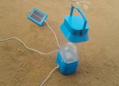 A blue and transparent solar lantern sits outside on the ground, connected to a tiny panel that is charging during the day