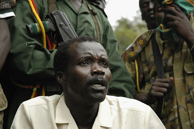 A man speaking while seated and flanked by two armed guards