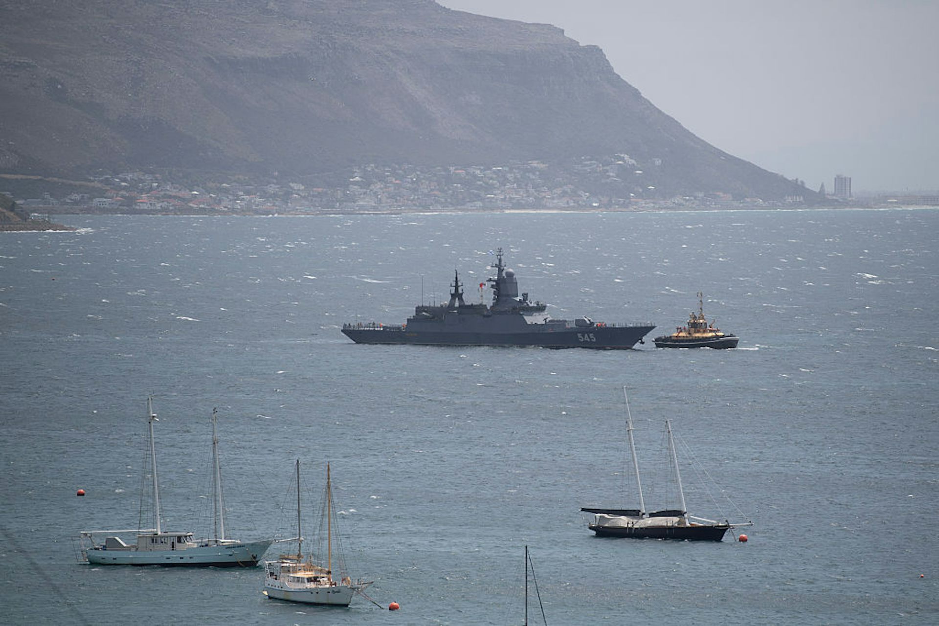 A large ship surrounded by four smaller boats in the middle of a vast body of water with a mountain in the background.
