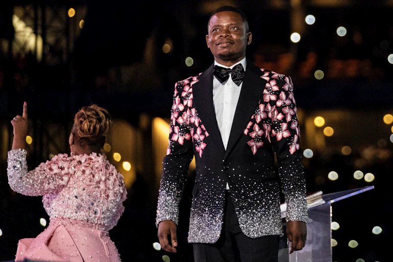 An African man stands on a stage in an elaborate suit with pink flowers on it, behind him a woman in pink raises a finger in the air, her back to him.