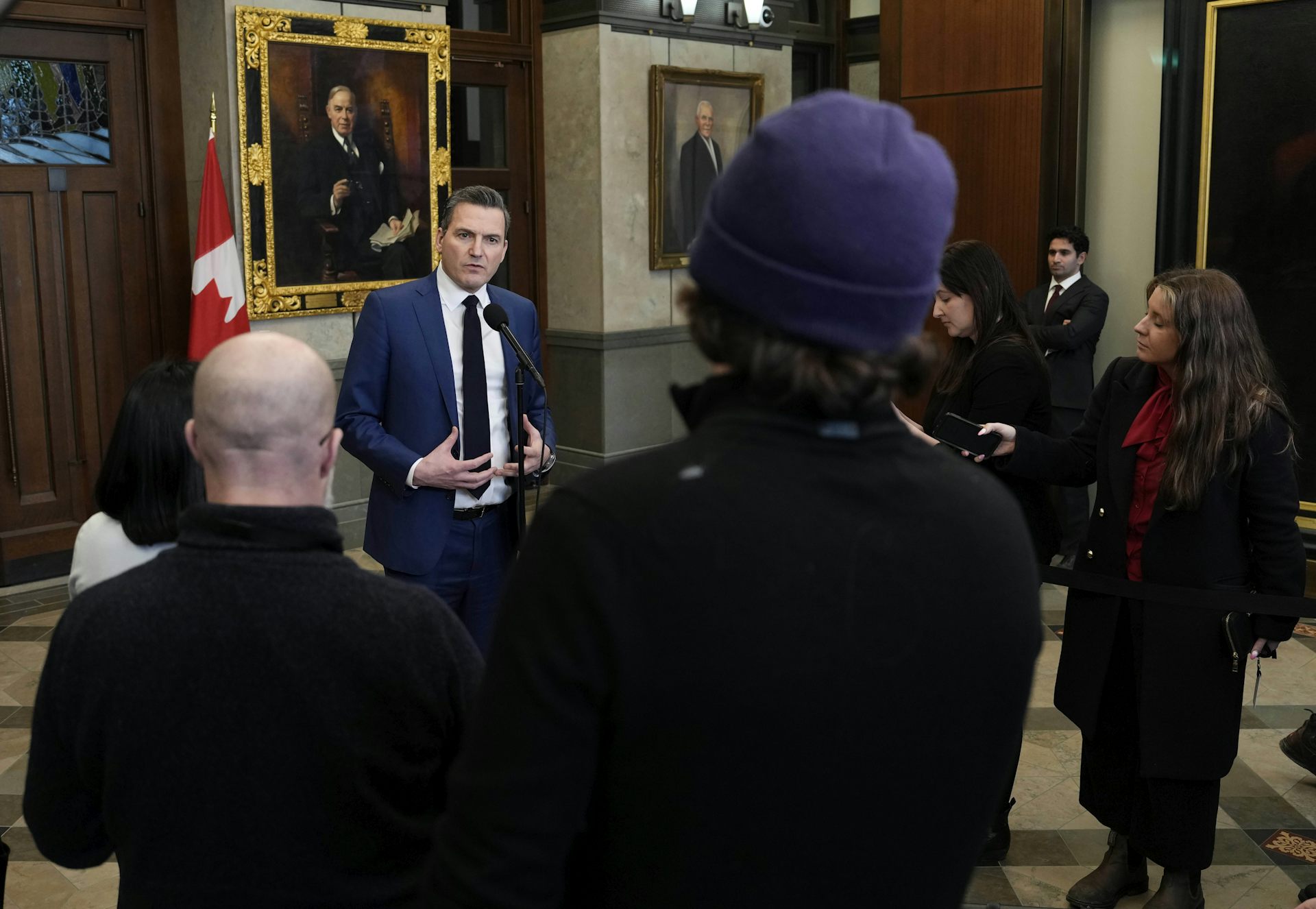 A man in a dark suit speaking in a large space, with other people seen from behind