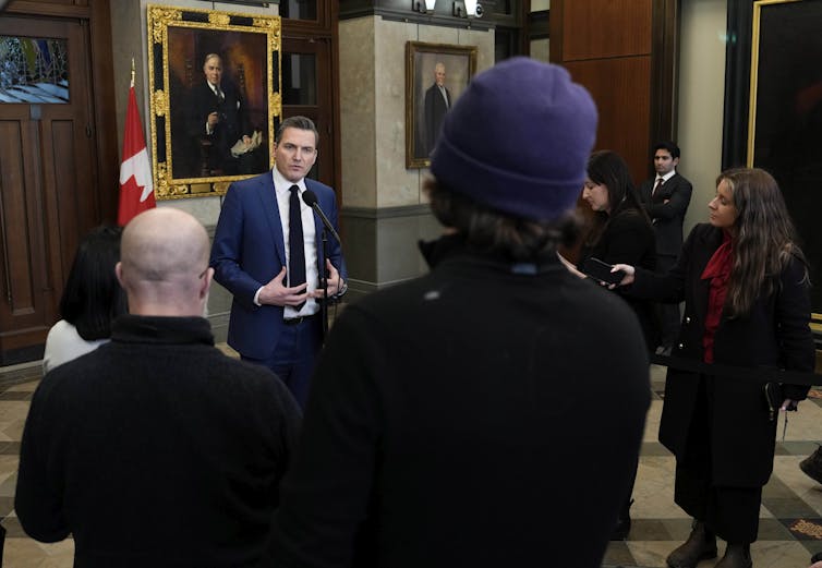 Un hombre con traje oscuro habla en un gran espacio, mientras se ve a otras personas desde atrás.
