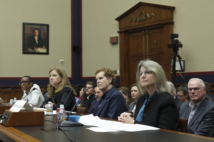 A row of four women dressed formally are seen sitting at a table together.