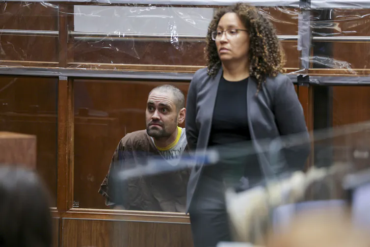In a courtroom, a tattooed man sits while his attorney stands beside him.