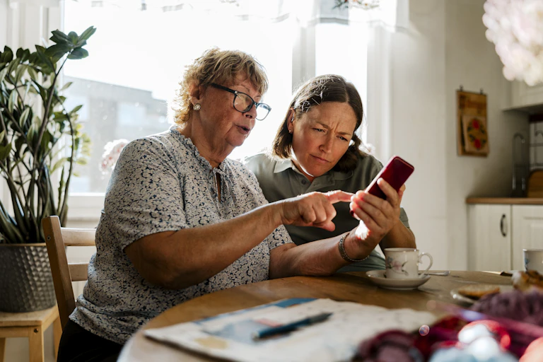 an older woman sitting at a table points to the screen of the smartphone she's holding as a younger woman looks where she's pointing