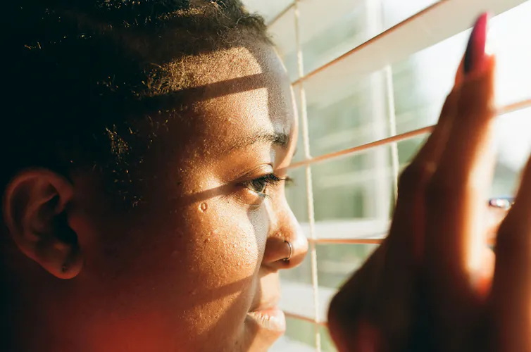 A Black woman looks out the window into sunshine through the shades of blinds.