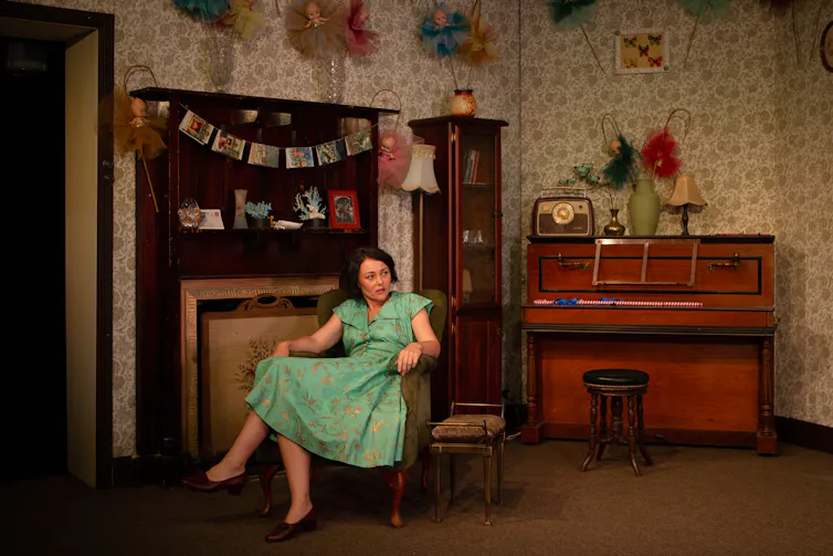 Production image: Olive sits in a chair, kewpie dolls on the walls.
