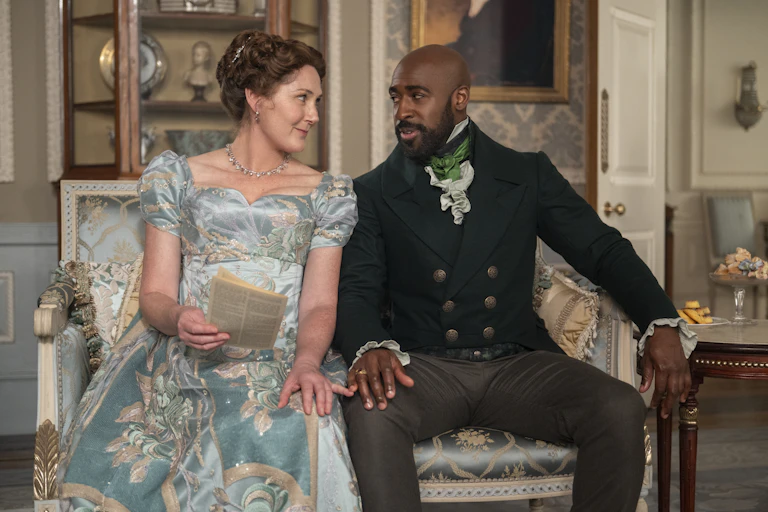 A couple in Regency-era clothing sit close together on a blue sofa, holding hands and gazing at each other.