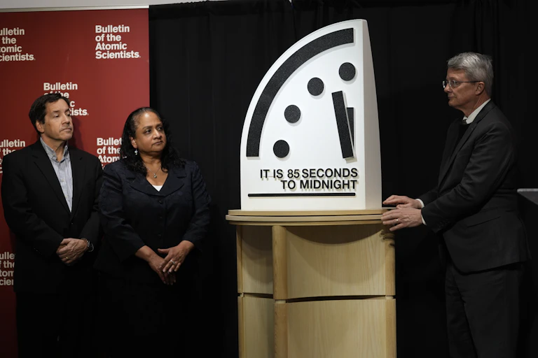 Two men and a woman stand next to a display that shows a portion of a clock and reads 'It is 85 seconds to midnight.'