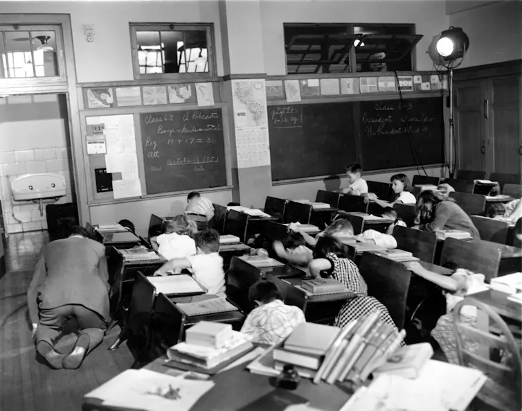 A black and white photo of schoolchildren and their teacher crouching on the floor of their classroom.
