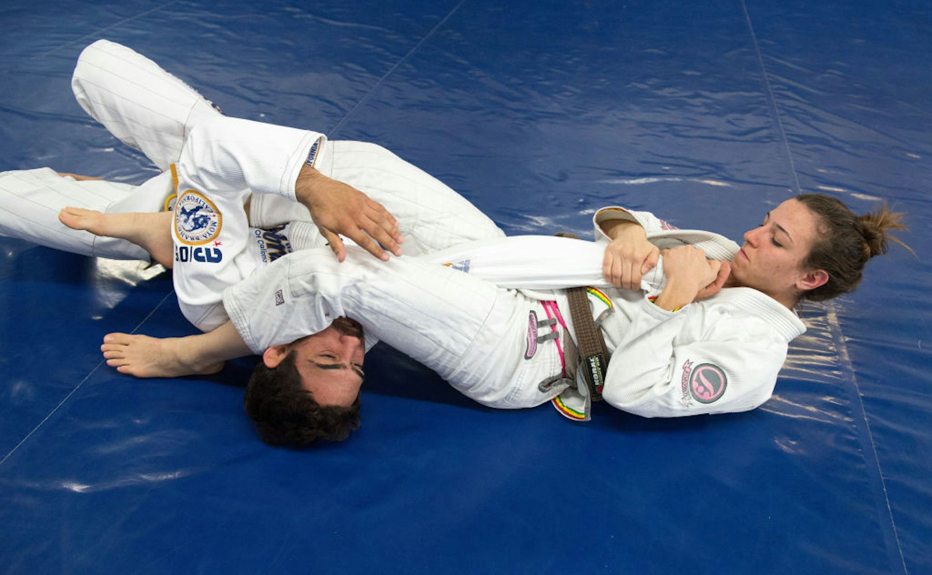 A young woman in a white martial arts suit performs an arm bar on a young man as the two grapple on a blue mat.