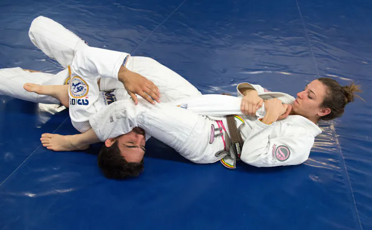 A young woman in a white martial arts suit performs an arm bar on a young man as the two grapple on a blue mat.