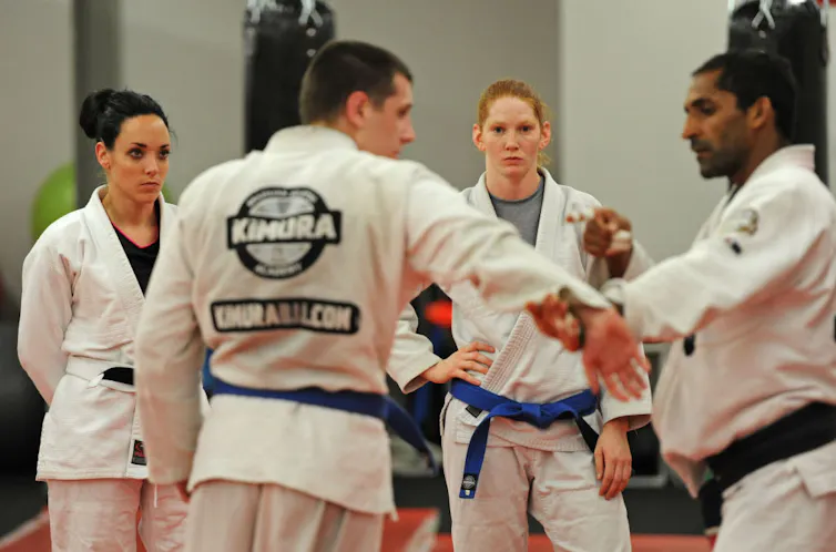 Two young women wearing white martial arts suits watch a young man in a white martial art suits hold the arm of another young man in a white martial arts suit.