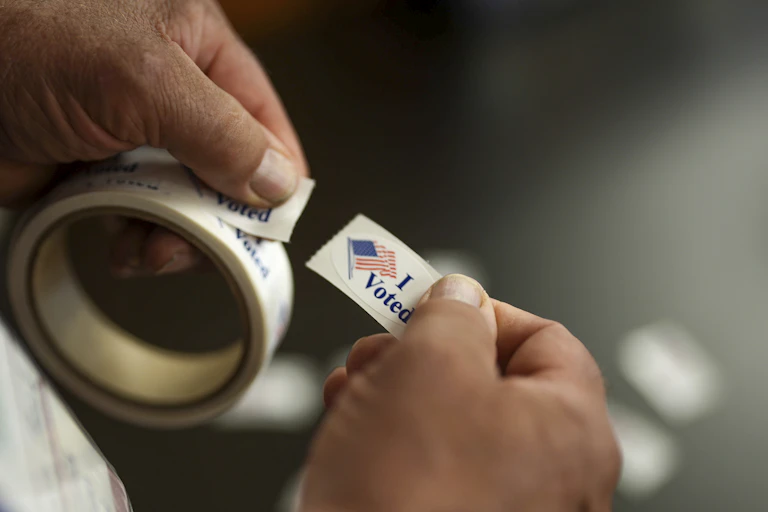hands are seen holding a roll of i voted stickers in the left and a single one in the right
