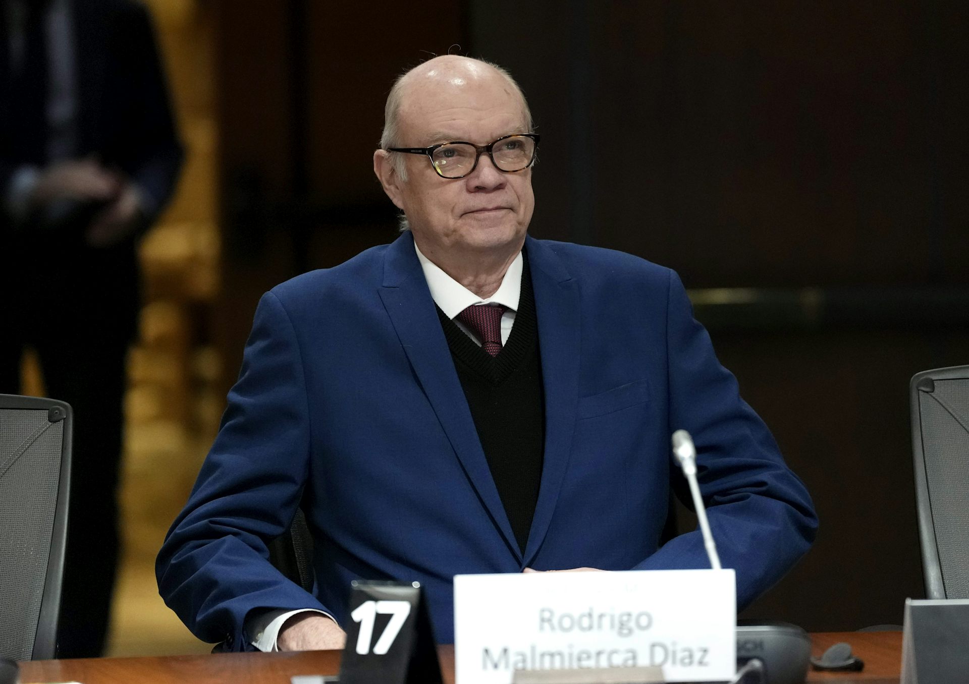 Un anciano calvo con traje azul y gafas se sienta en un escritorio con un micrófono.