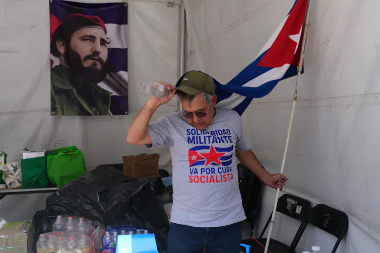 a man wearing a shirt with a cuban flag and holding the flag on a poll stands in a tent. A photo of Fidel Castro hangs behind him