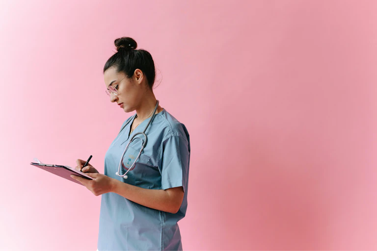 A young doctor writes notes on a clipboard, in front of a pink background.