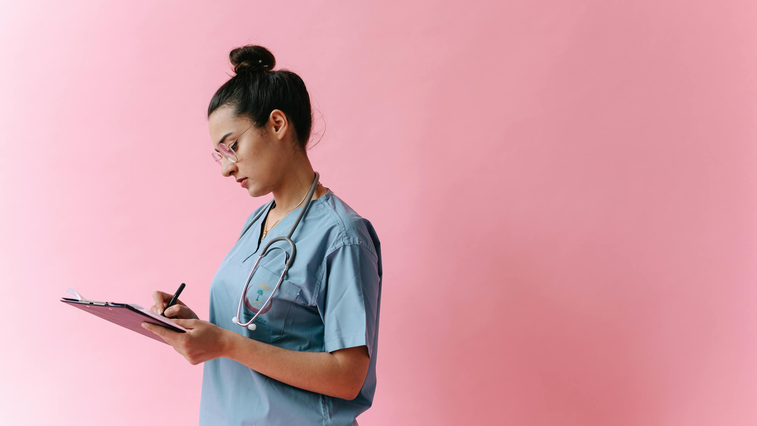 A young doctor writes notes on a clipboard, in front of a pink background.