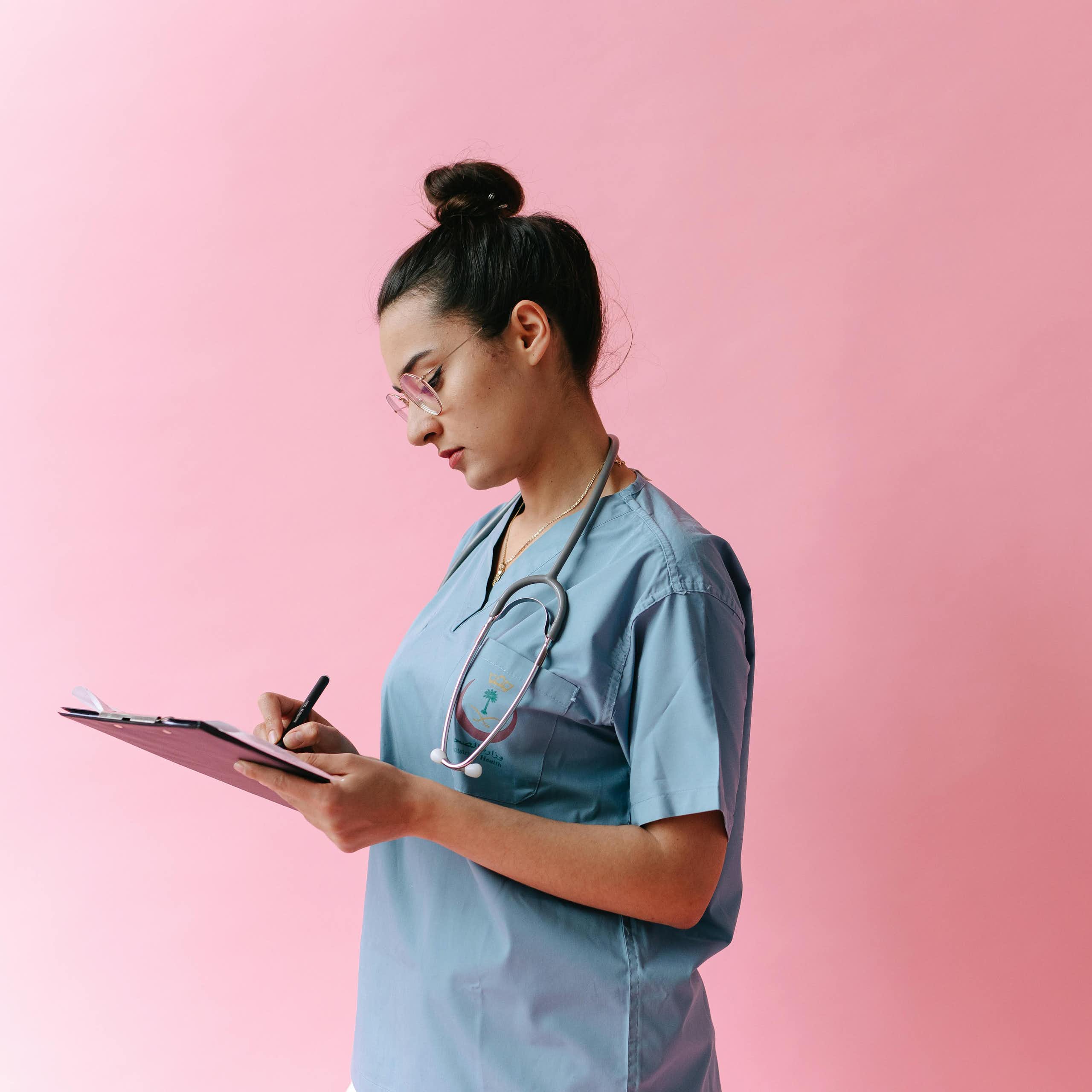 A young doctor writes notes on a clipboard, in front of a pink background.