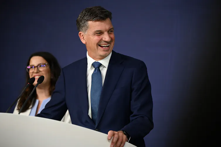 A man in front of a blue backdrop at a lectern smiles and looks to the side