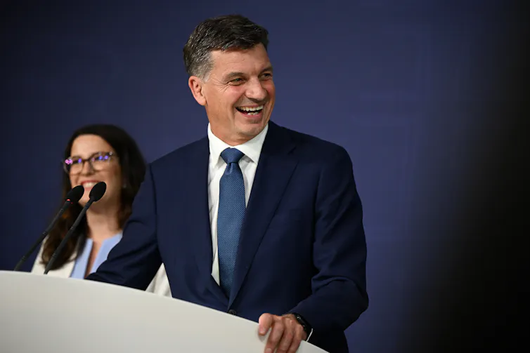 A man in front of a blue backdrop at a lectern smiles and looks to the side