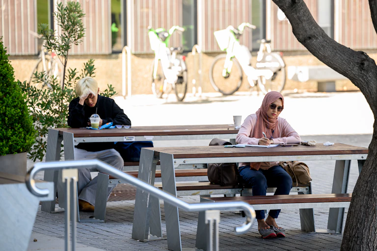 Two young people sit at outside benches. One has his head in his hands.