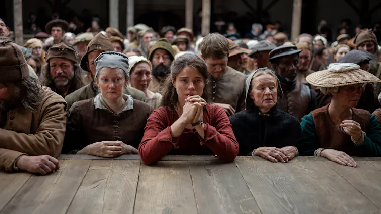 Production still: teary-eyed woman with hands clasped together looks directly at the camera