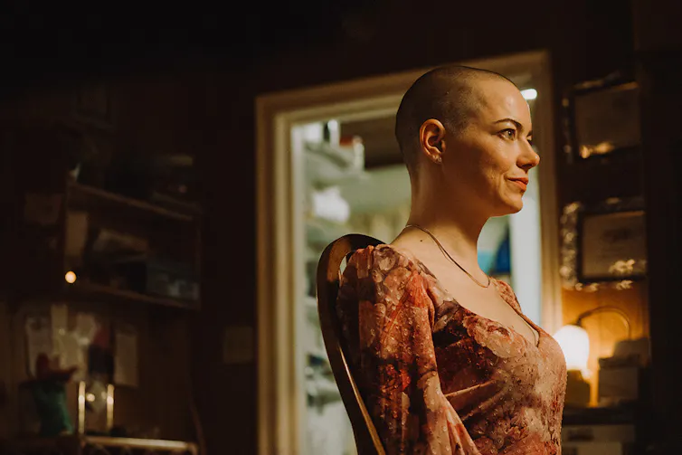 Production still: mid-shot of a bald woman seated on a chair facing the right, smiling off-camera.