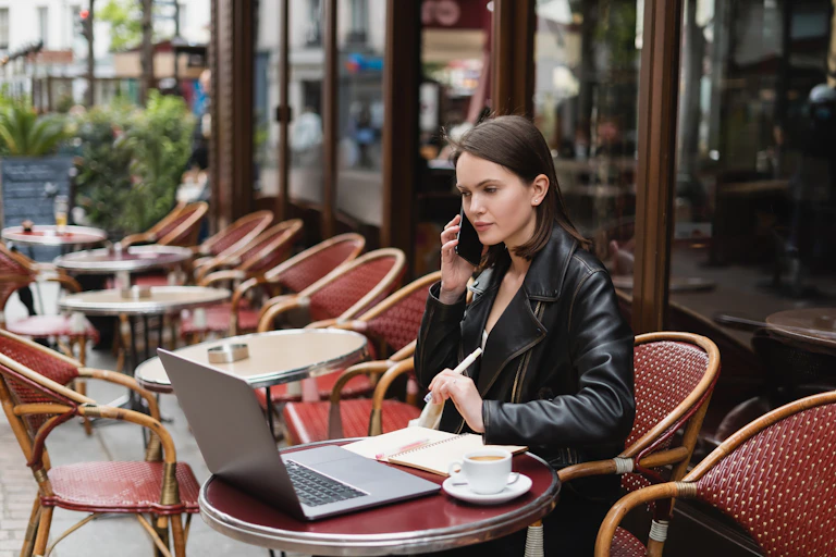 A woman makes a phone call, sitting at a cafe table where she is working on her laptop.