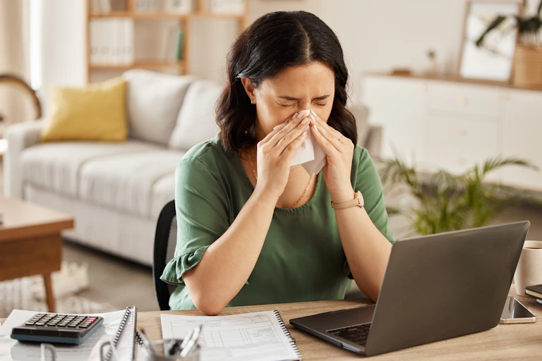 A woman sitting in front of her laptop blows her nose into a tissue.